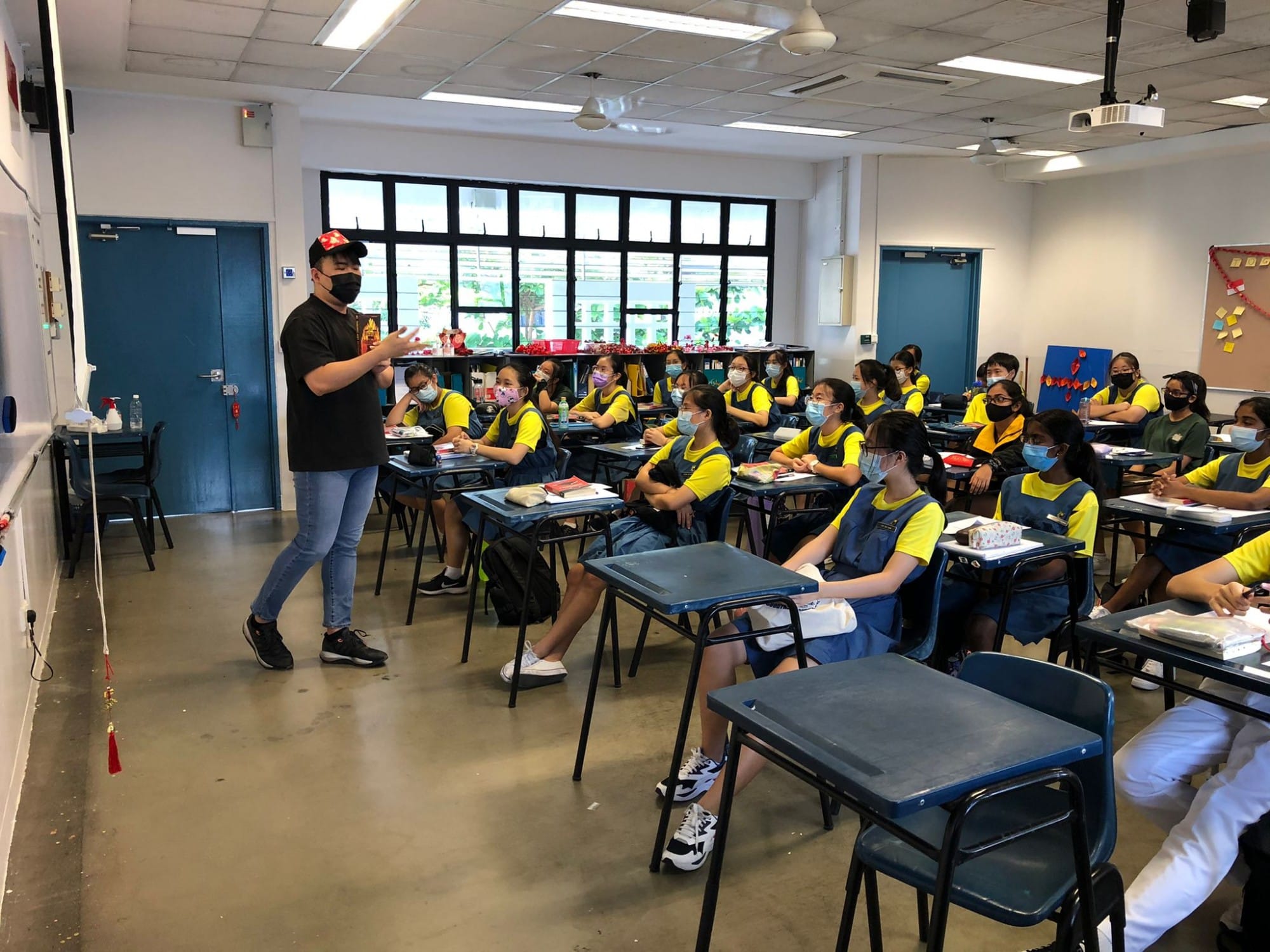 A masked teacher in black stands before rows of students in yellow uniforms wearing masks in a classroom.